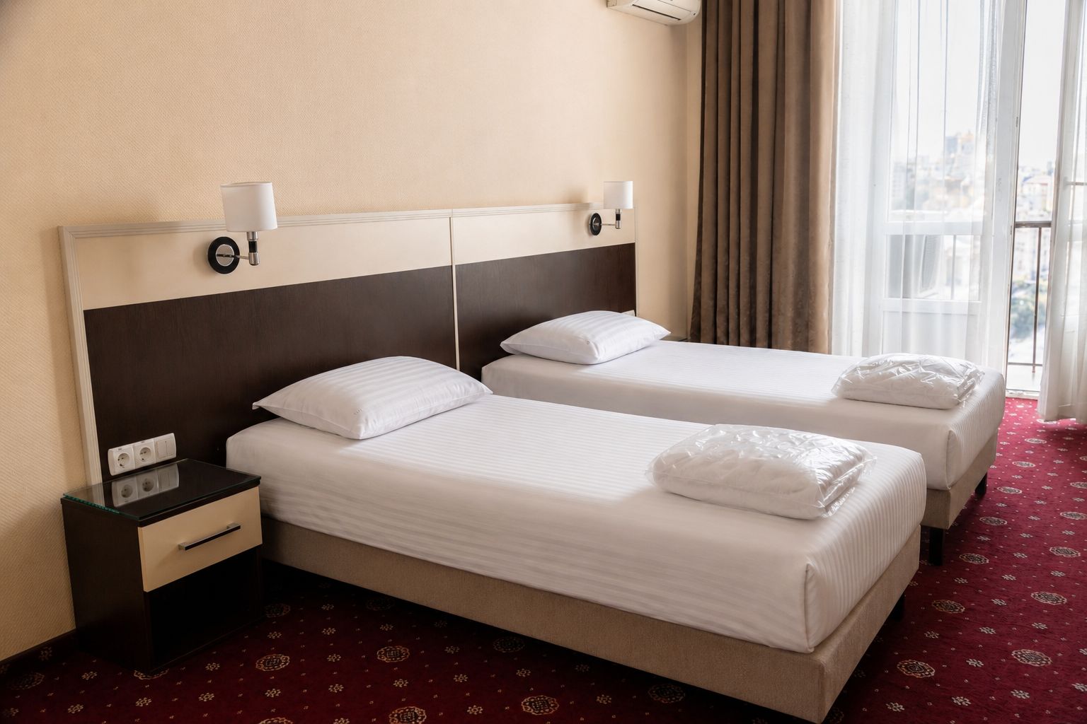 A hotel room with two beds, a nightstand, a lamp, and a window with curtains.
