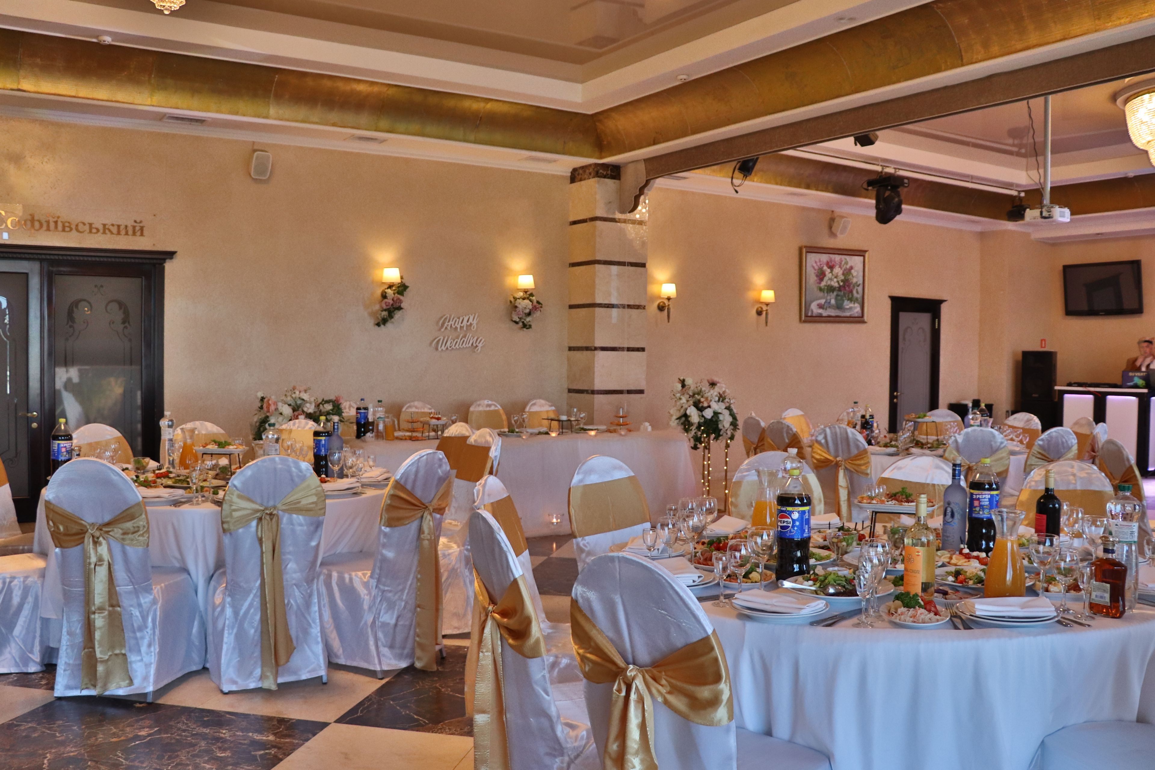 A banquet hall with round tables set for a wedding reception, featuring white tablecloths and chairs with gold sashes.