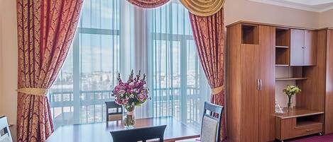 Une chambre d’hôtel équipée d’une table à manger, de chaises, d’un vase de fleurs, d’une armoire en bois et de grandes fenêtres avec des rideaux.