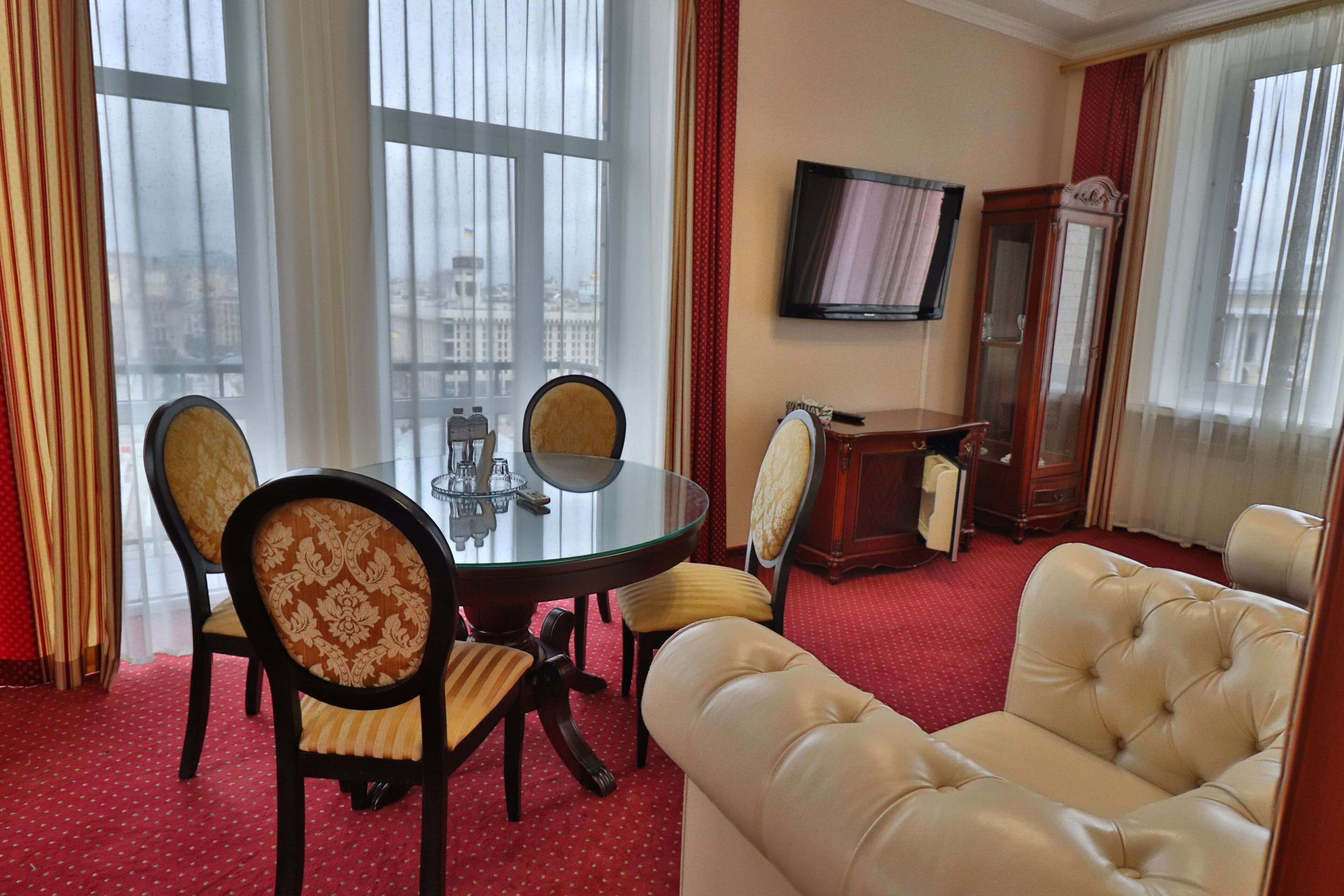 Habitación de hotel con una mesa de comedor, sillas, un sofá, un televisor y un amplio ventanal con cortinas.