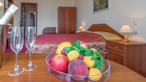 Una habitación de hotel con una mesa de madera, una cama con una manta roja y un frutero con manzanas y melocotones.