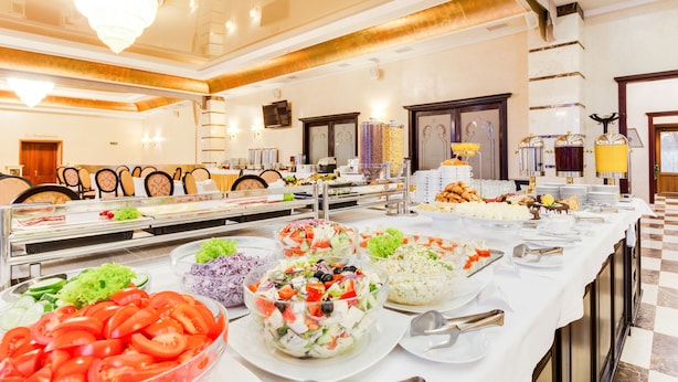 Un bufé con diversos alimentos, incluyendo ensaladas y platos de arroz, en una sala bien iluminada con candelabro y suelo a cuadros.