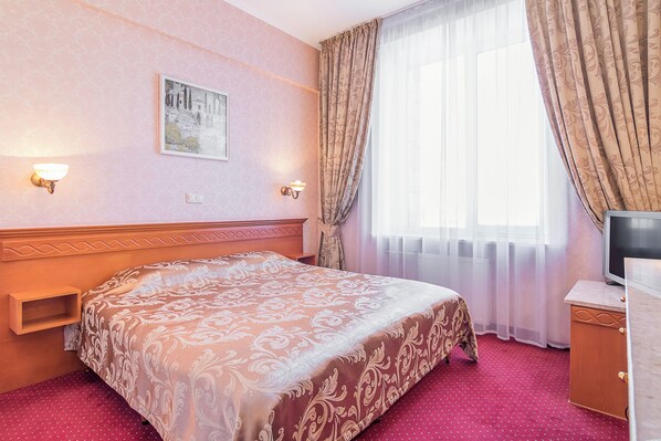 A hotel room with a large bed, a television on a wooden cabinet, a wall-mounted light, and a window with curtains.