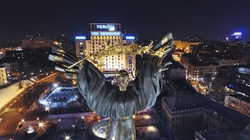 Vista noturna de uma paisagem urbana, com uma estátua em destaque ao primeiro plano e um hotel com letreiros iluminados ao fundo.