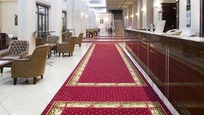 Un vestíbulo de hotel con alfombra roja, zona de estar, mostrador de recepción y una escalera que sube a otro piso.