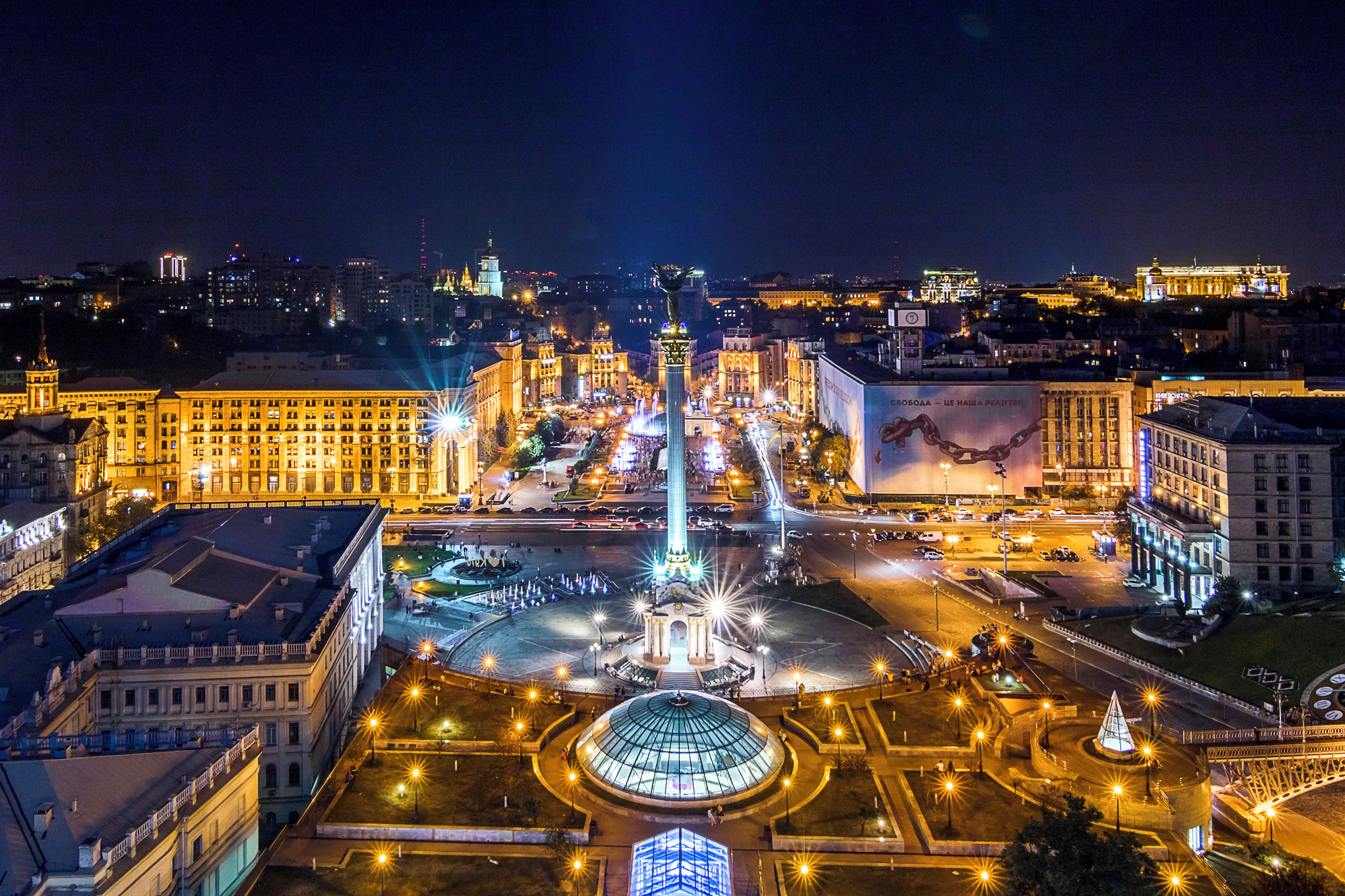 A city square with a central monument, surrounded by buildings with illuminated facades.