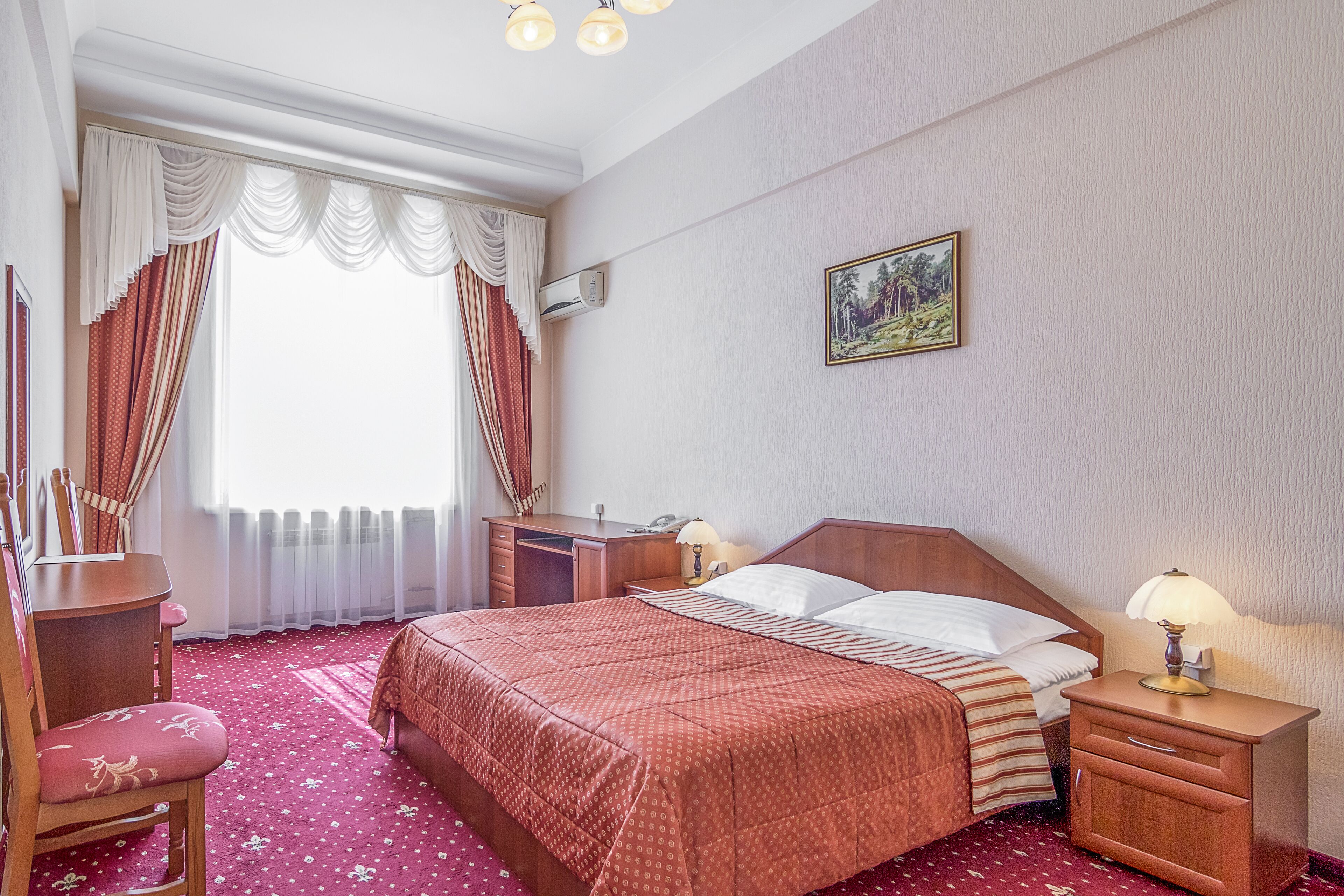 Habitació d'hotel amb un llit, escriptori, cadira, tauleta de nit amb una làmpada i un quadre a la paret.