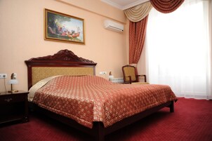 Una cama bien hecha con una colcha estampada, un cabecero de madera, una mesita de noche con una lámpara, una silla, un cuadro en la pared y una ventana con cortinas.