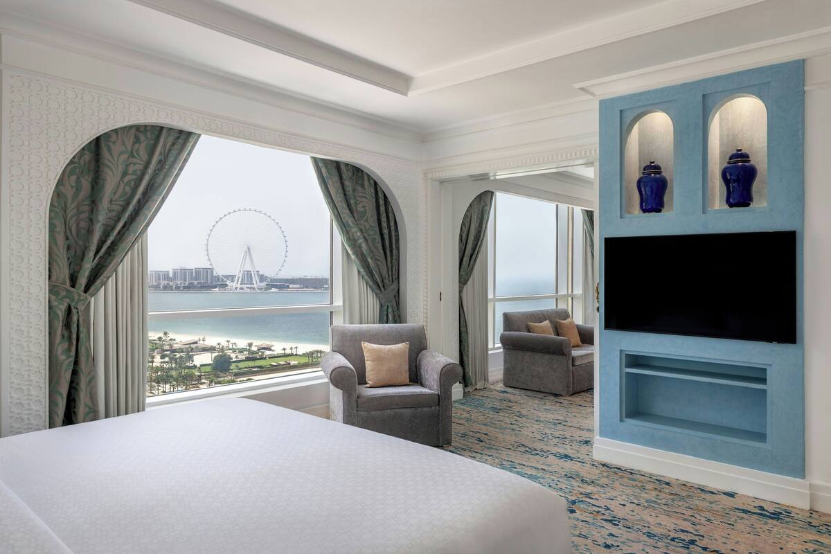 Una camera d'albergo con un letto grande, due poltrone, una TV e vista su una ruota panoramica.