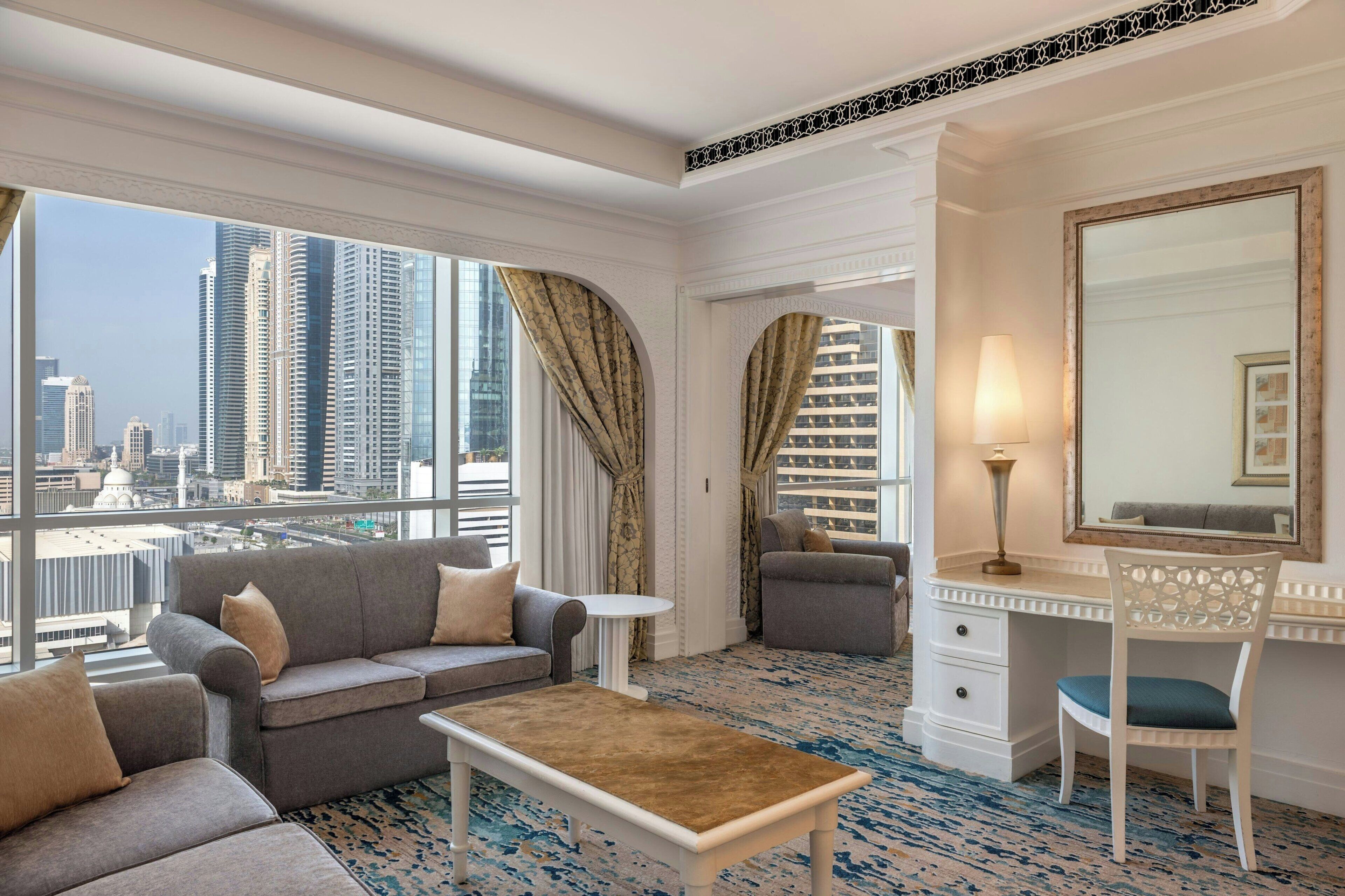 Une chambre d’hôtel moderne avec vue sur la ville, comprenant un canapé, un fauteuil, un bureau et un grand miroir.