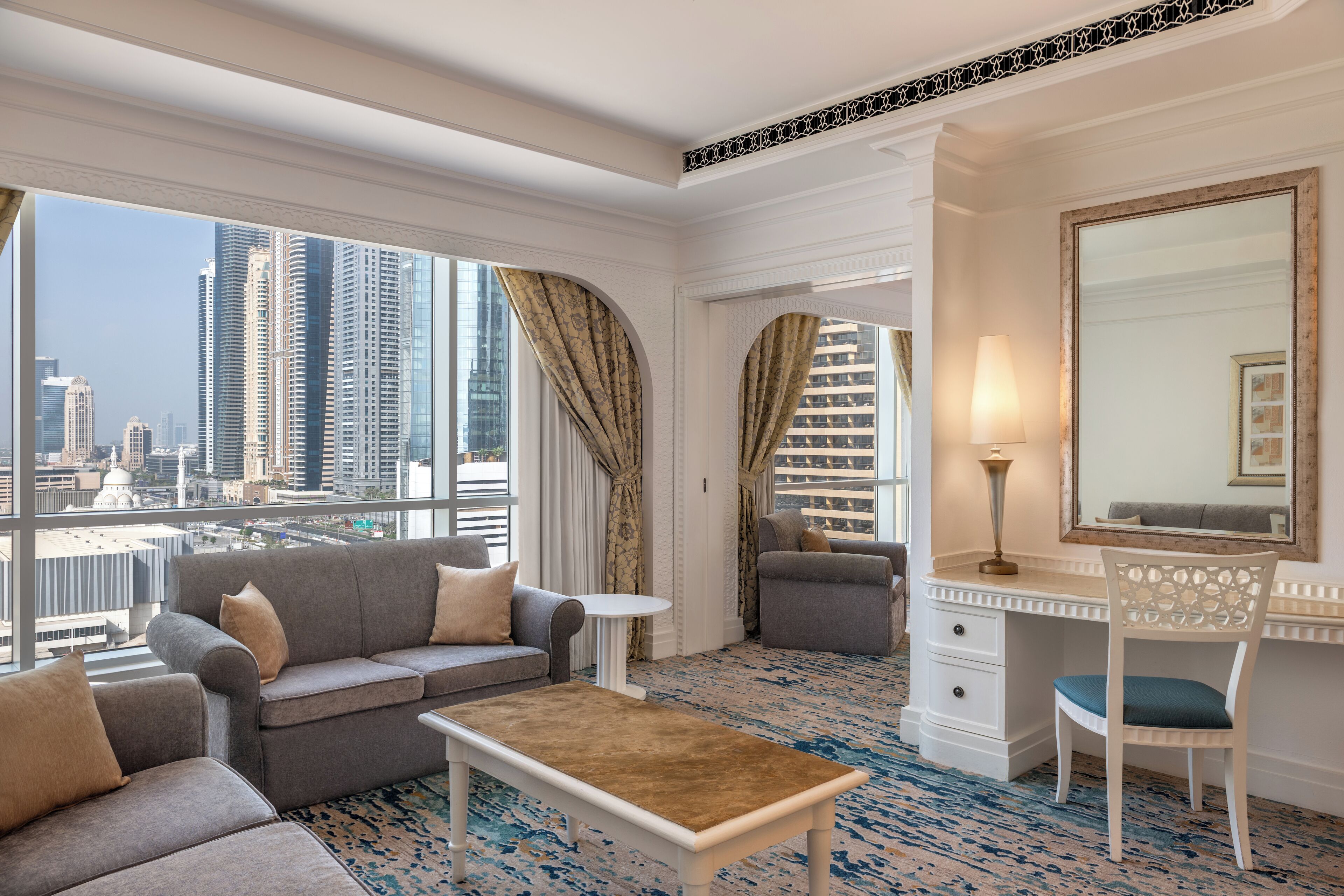 A hotel room with a large window offering a city view, a sofa, a chair, a desk, and a mirror.