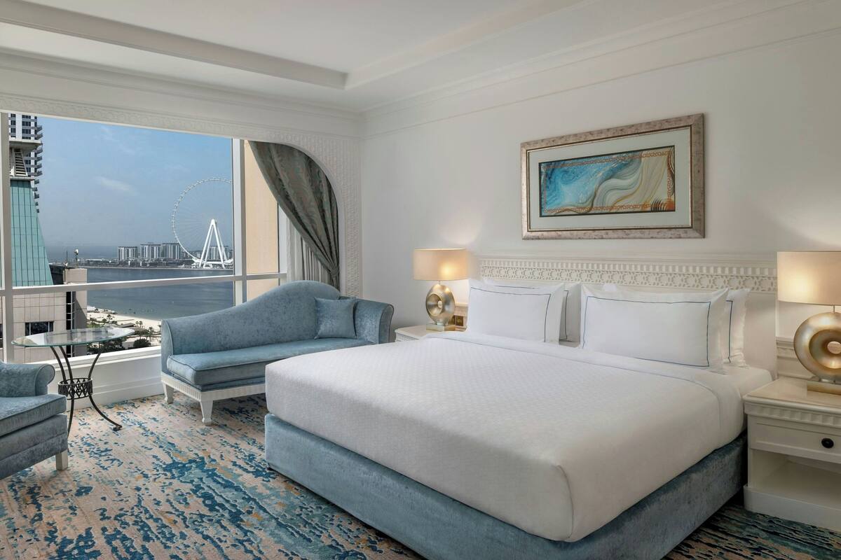 Una camera d'albergo con un letto grande, due poltrone, un tavolino e vista su una ruota panoramica e degli edifici.