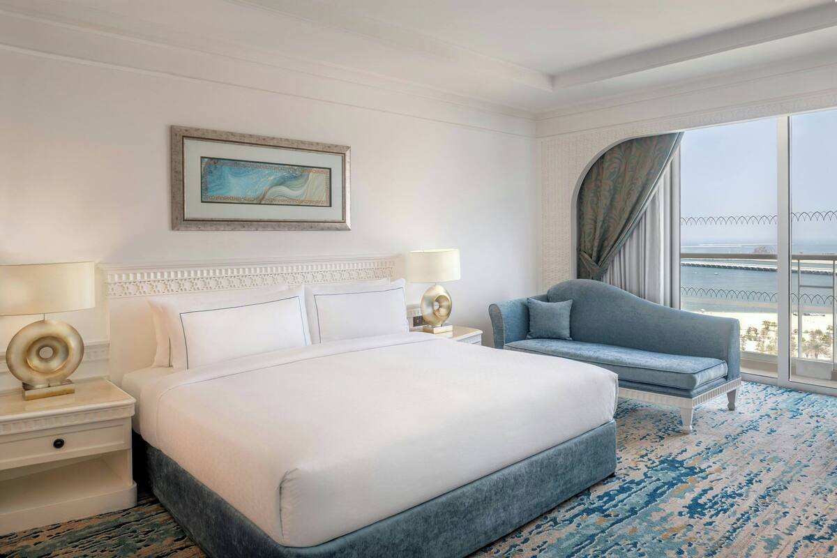 Camera d'albergo con un letto grande, un divano blu, un comodino con una lampada e una finestra vista mare.