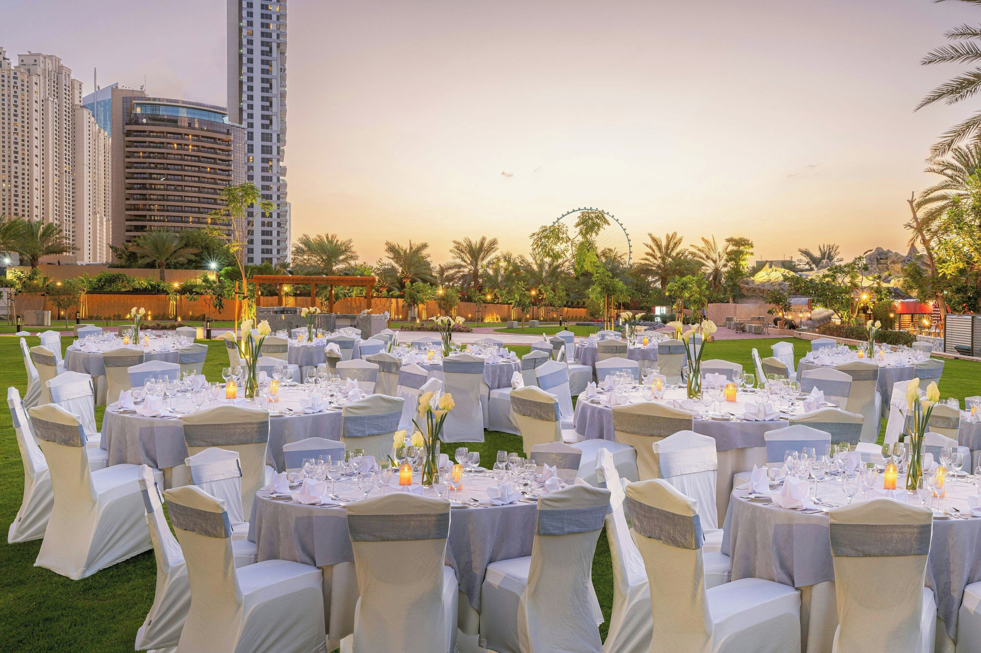 An outdoor event space with round tables set for dining, surrounded by chairs with white and grey covers, and a backdrop of palm trees and buildings.