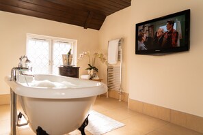A bathroom with a freestanding bathtub, a television mounted on the wall, and a window with blinds. -  The Kingfisher Pub & Hotel (Bedford)