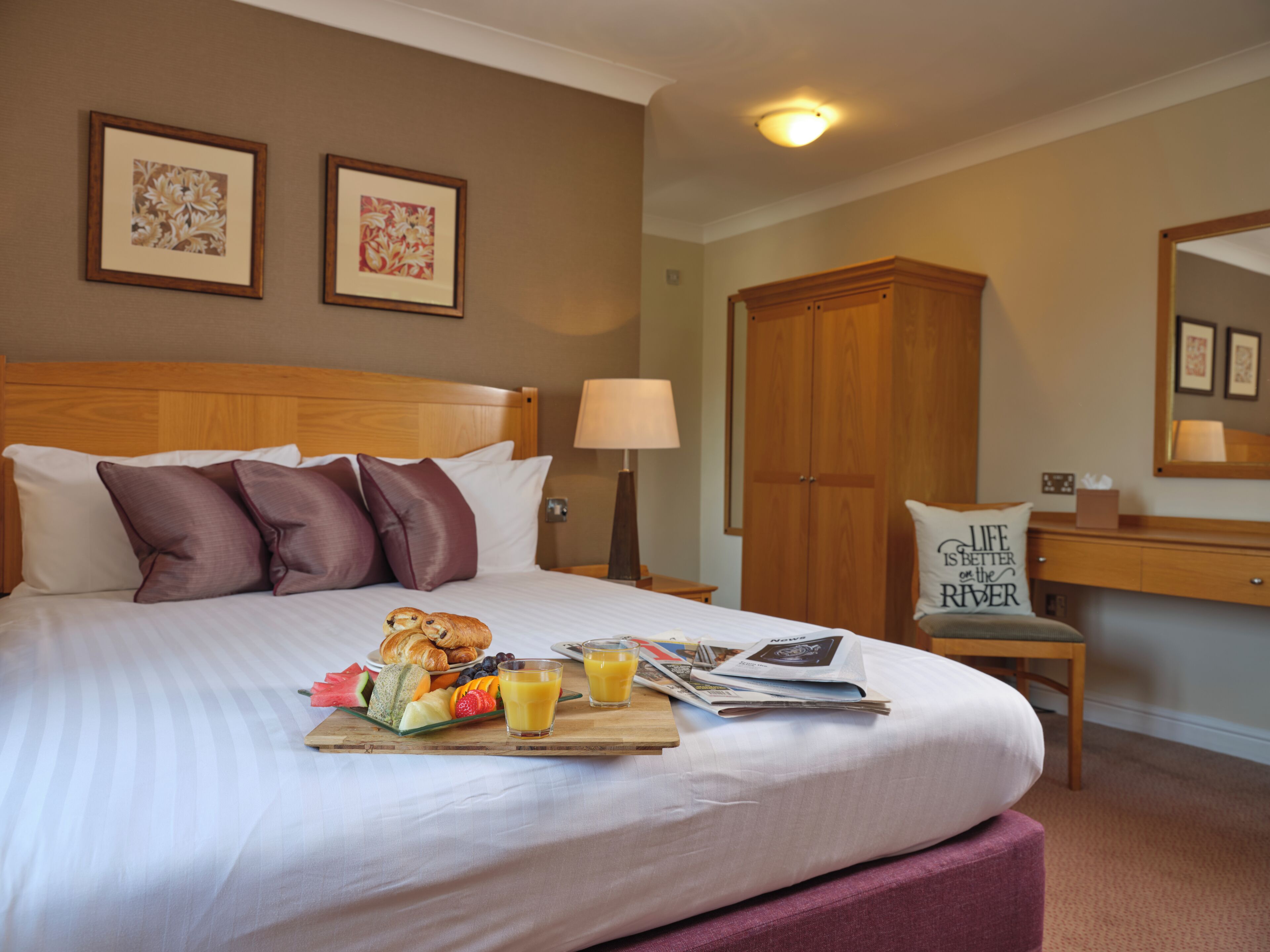 Een hotelkamer met een keurig opgemaakt bed, een ontbijtkarretje met fruit en sap, een houten garderobe en een bureau met een lamp.
