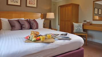A hotel room with a neatly made bed, a breakfast tray with fruits and juice, a wooden wardrobe, and a desk with a lamp.