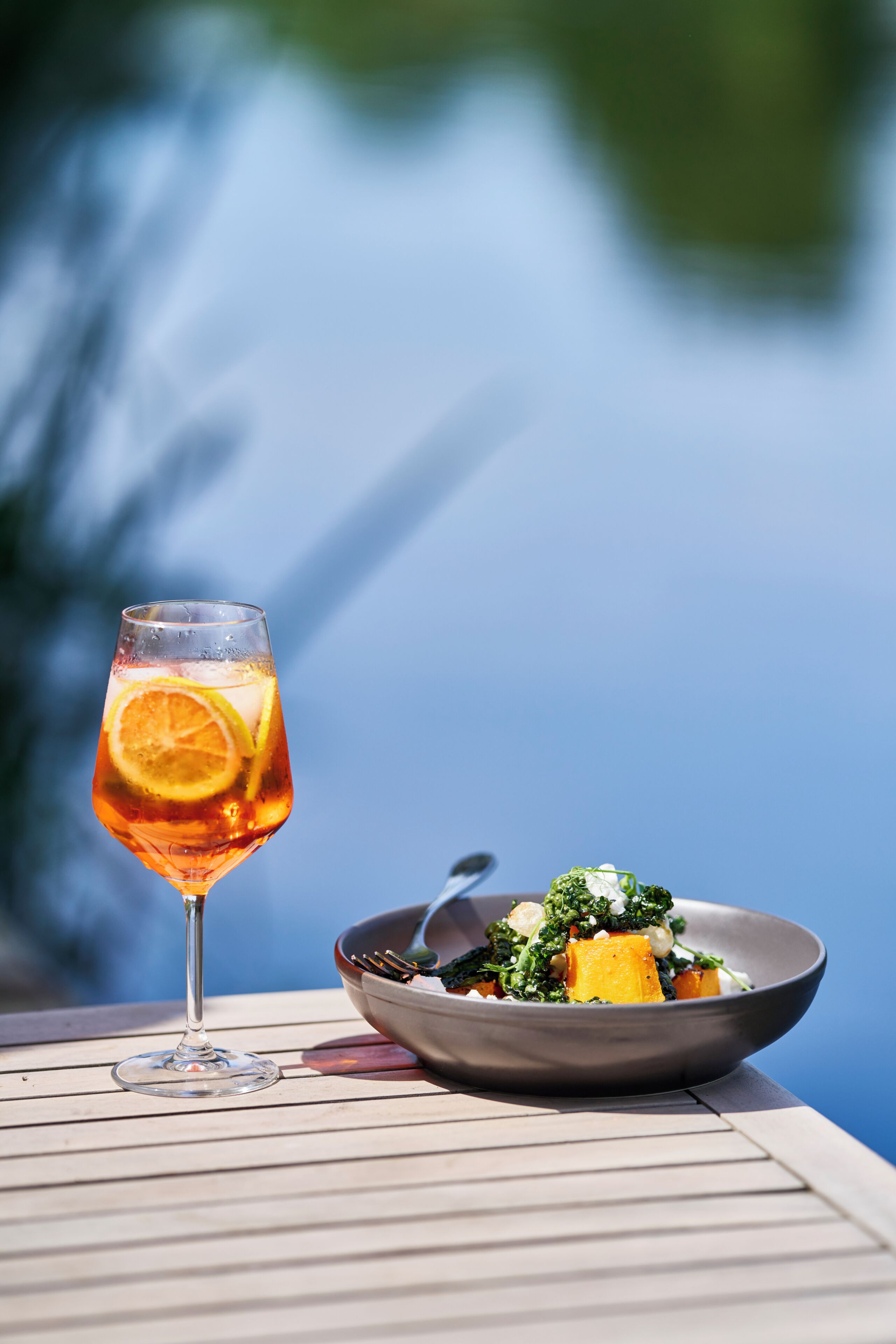 A glass of drink with a slice of orange on a table with a bowl of food beside it.