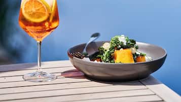 A glass of drink with a slice of orange on a table with a bowl of food beside it.