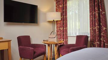 A hotel room with a bed, two armchairs, a small table with a lamp, a television, and patterned curtains.