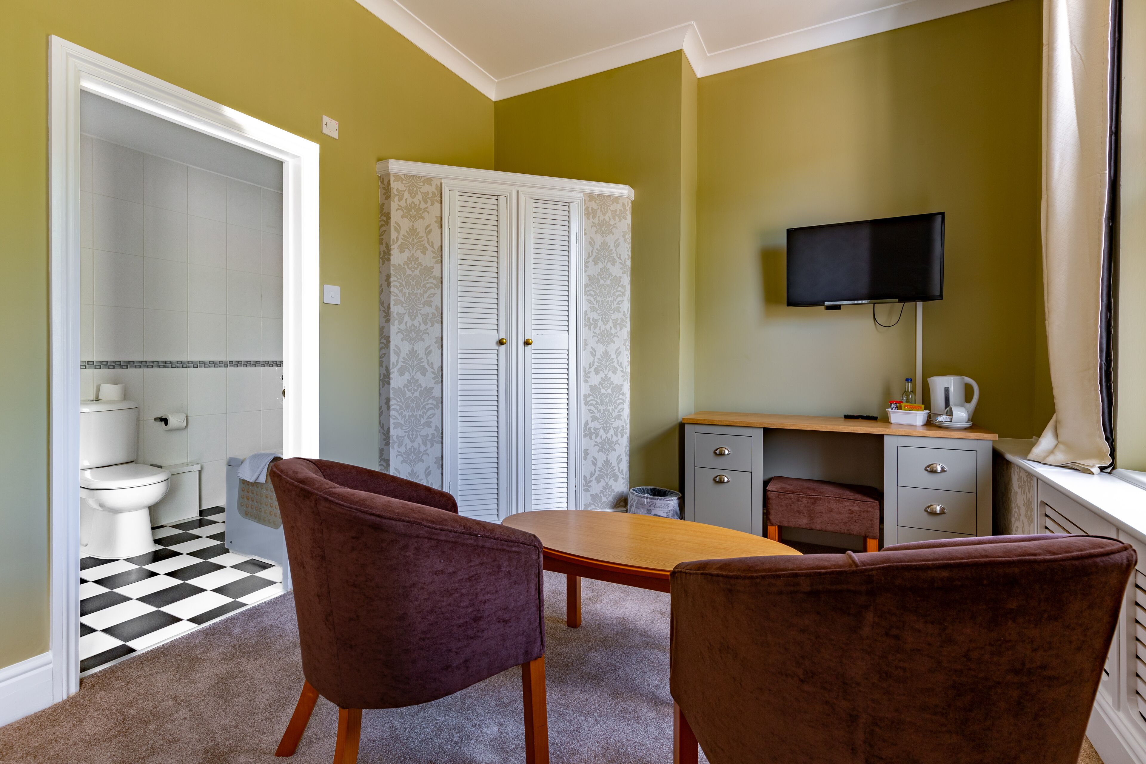 A room with a checkered floor, a wooden table, two chairs, a television, and a wardrobe.