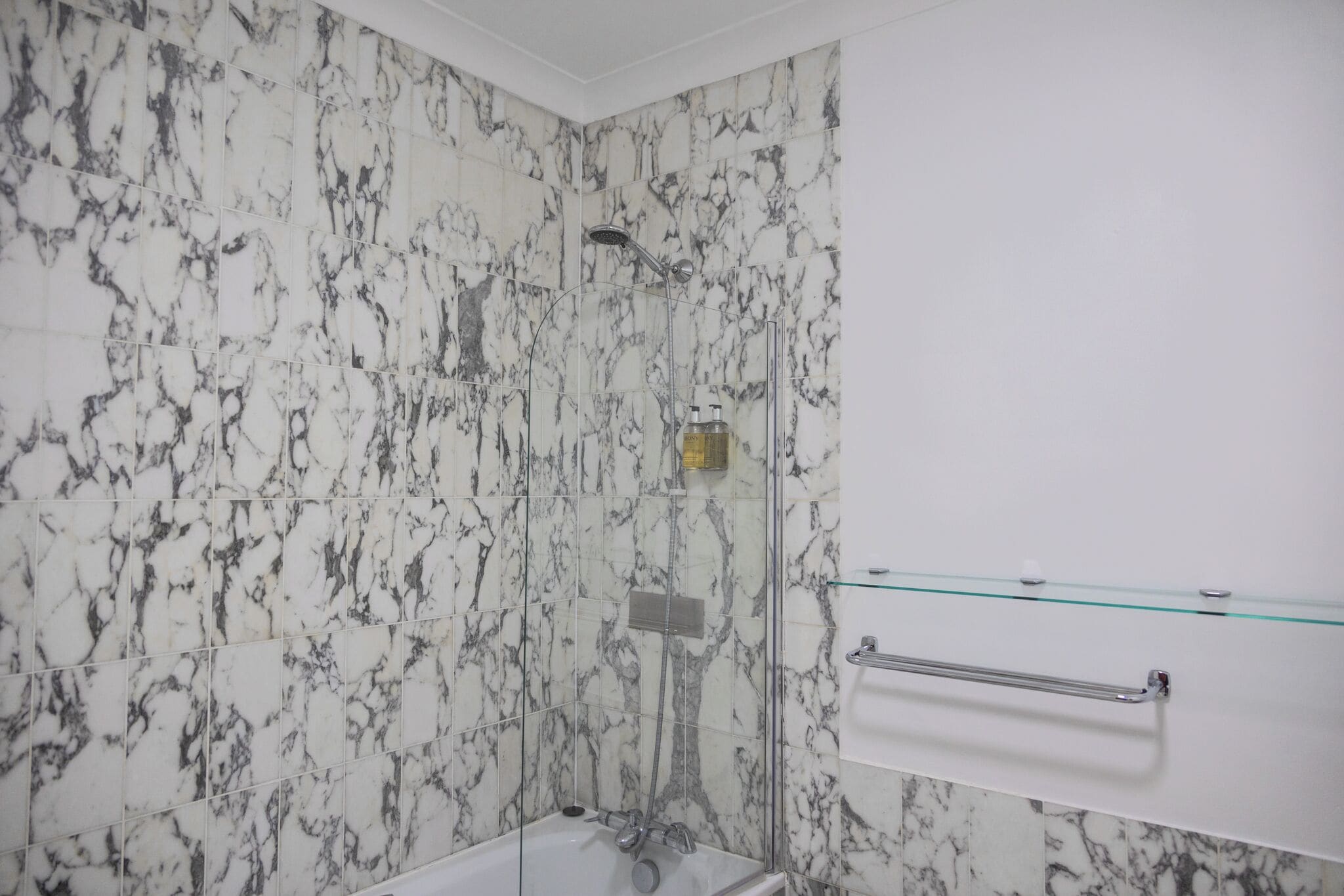 Un baño con paredes de mármol, cabina de ducha de vidrio y estante de vidrio con artículos de aseo.