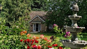Ein Backsteinhaus mit weißer Tür und Fenstern, umgeben von einem Garten mit einer Steinfunktion und verschiedenen Blumen.