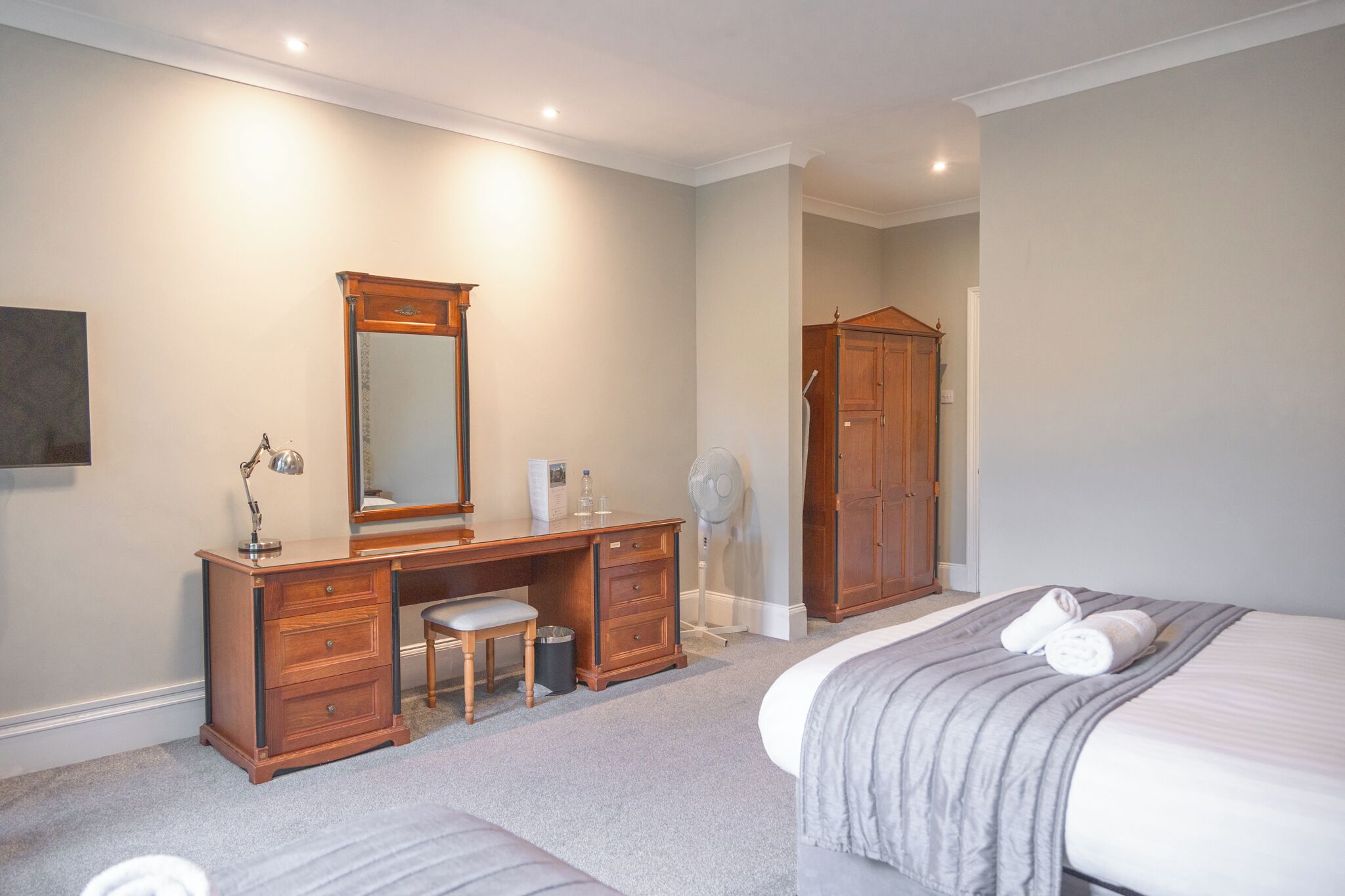 A bedroom with a bed, a wooden dresser, a desk, a chair, a mirror, a wardrobe, and a fan.