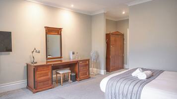 A bedroom with a bed, a wooden dresser, a desk, a chair, a mirror, a wardrobe, and a fan.