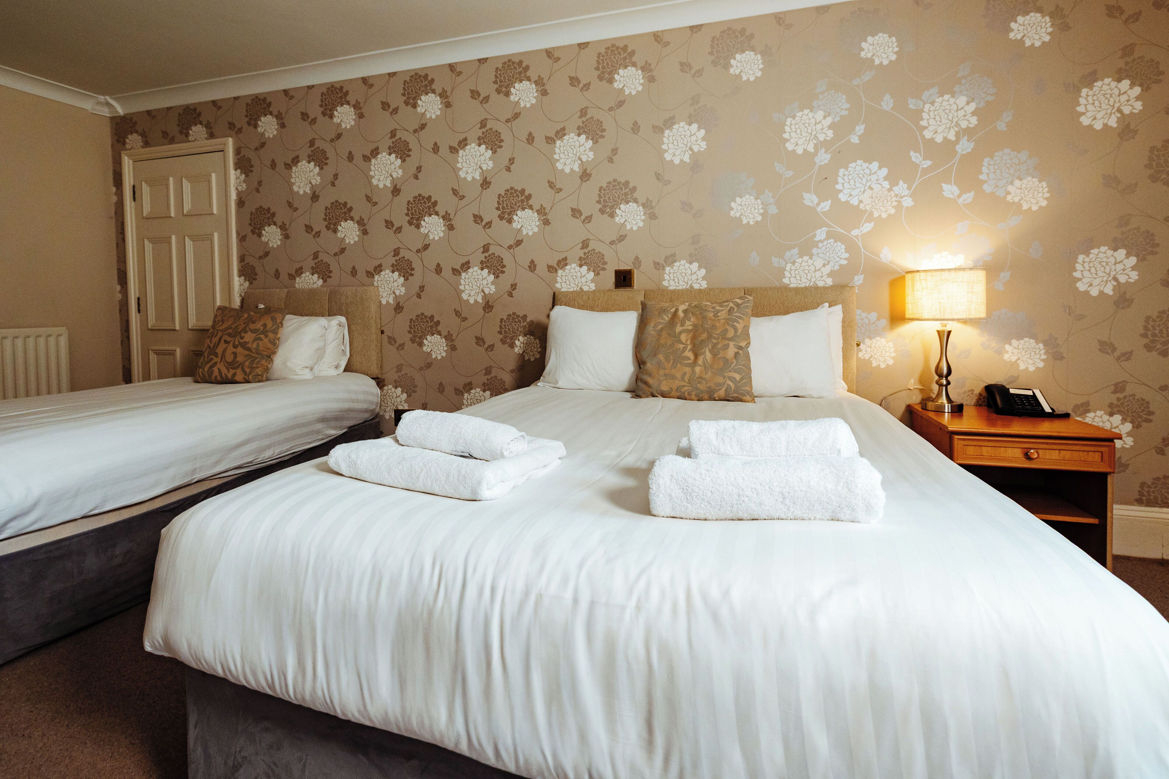 A hotel room with two beds, floral wallpaper, a bedside lamp, and a telephone on the nightstand.