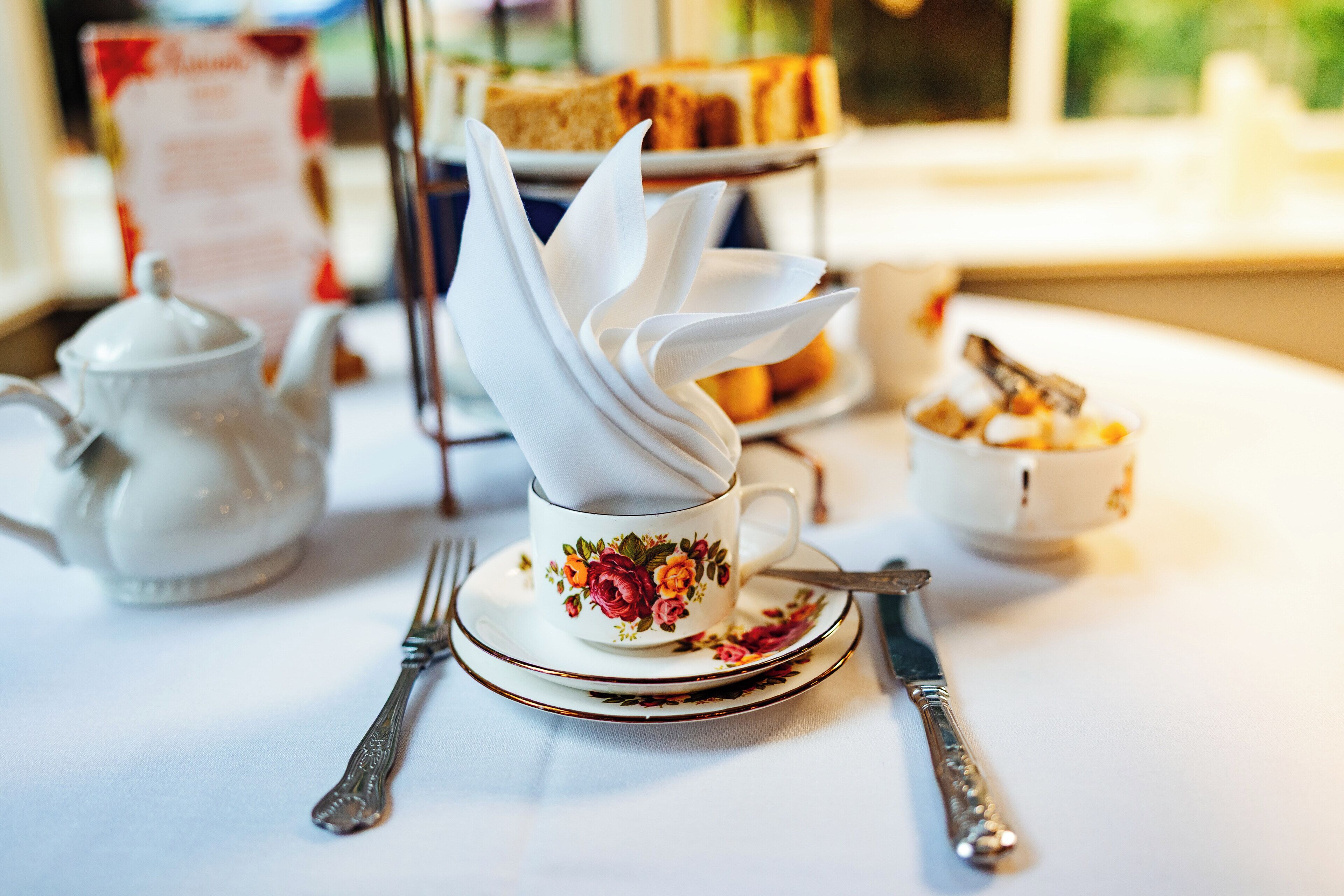 A tea set with a floral cup and saucer, a teapot, and a dessert display in the background.