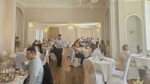 Ein stilvolles Esszimmer mit weißen Tischen und Stühlen, einem Kronleuchter und einem Kellner, der Gäste bedient.