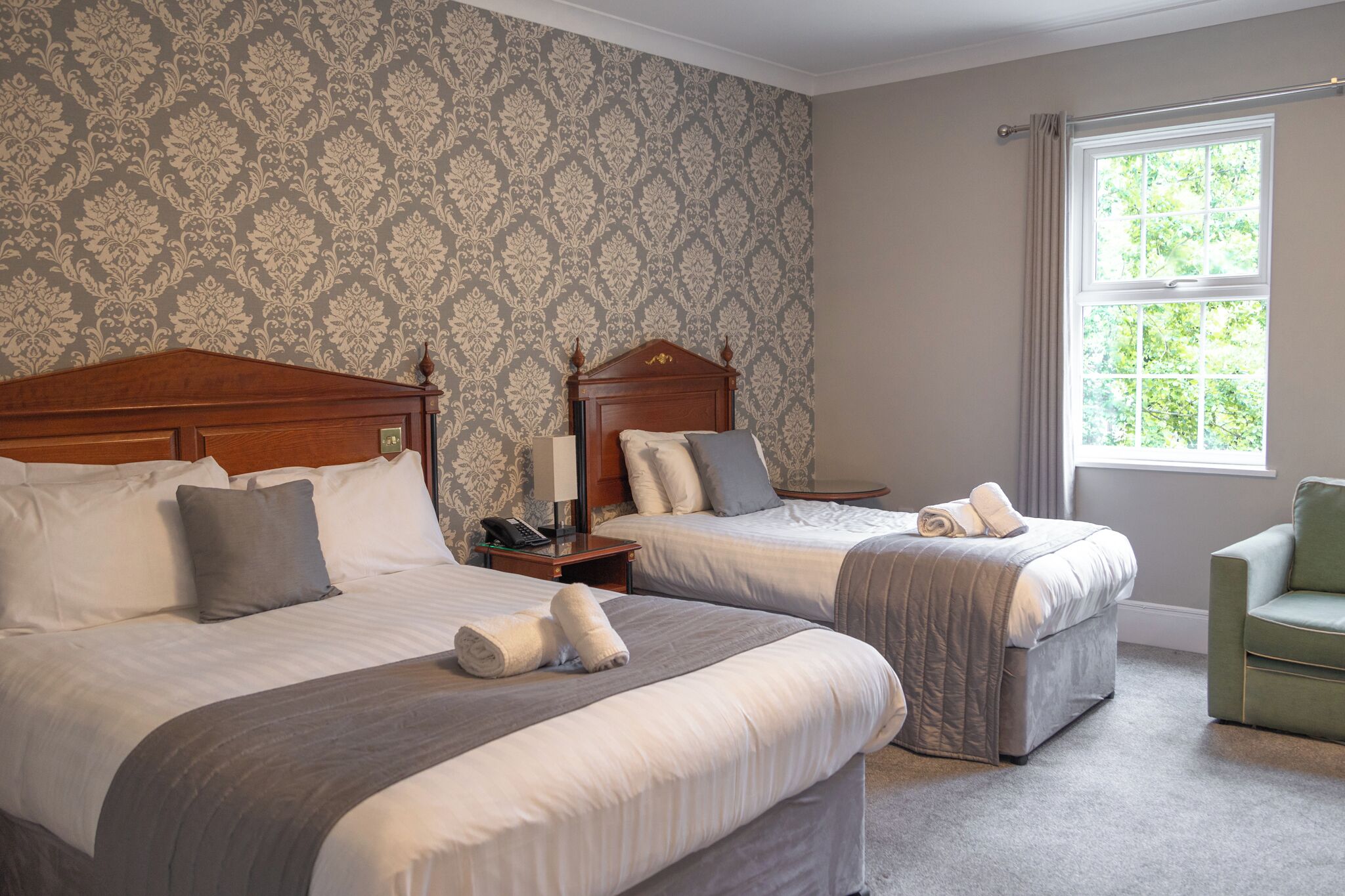 Een hotelkamer met twee bedden, een groene fauteuil en een raam met uitzicht op groen.