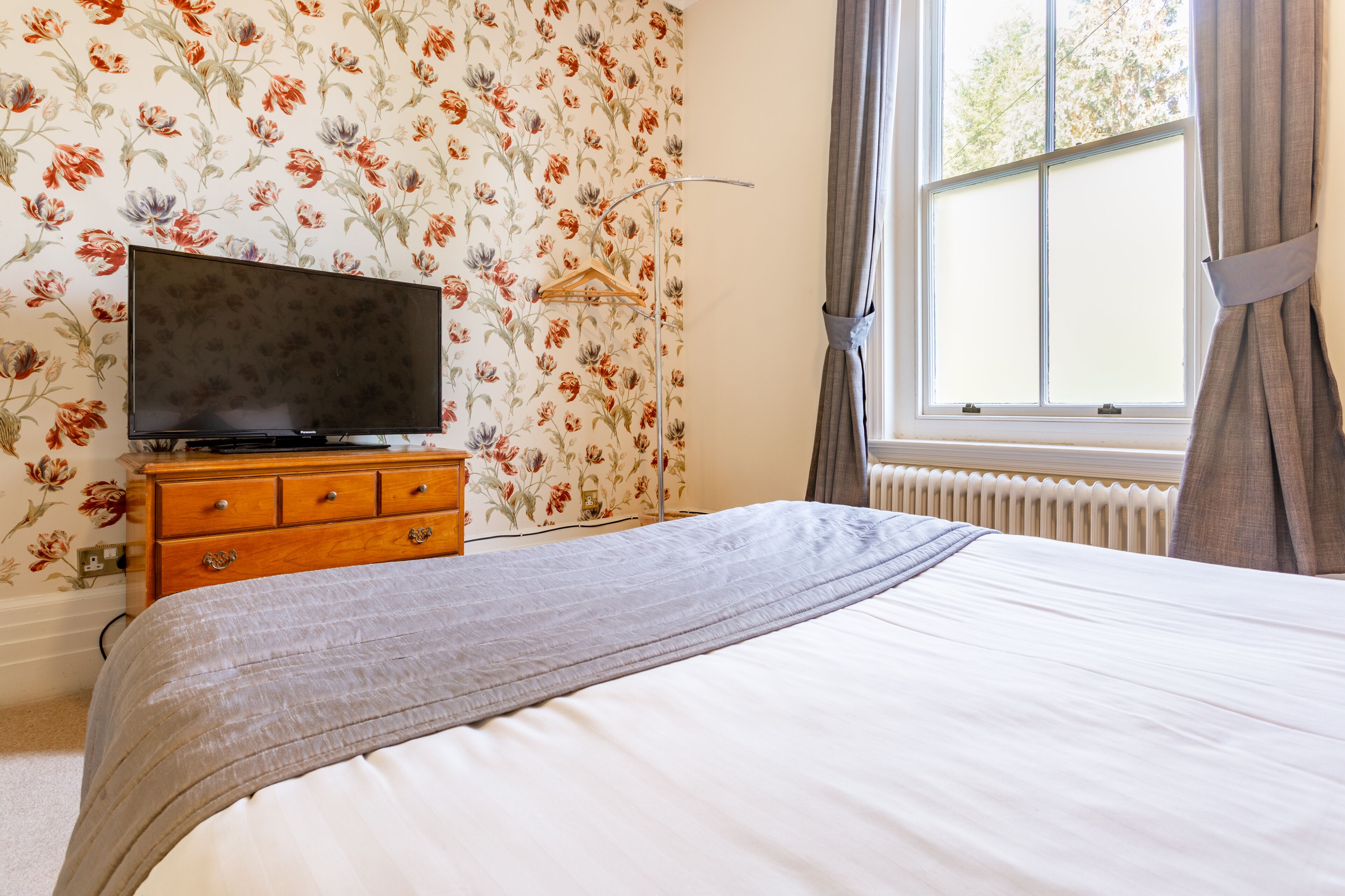 A bedroom with a bed, a dresser, a television on the dresser, floral wallpaper, a window with curtains, and a radiator.
