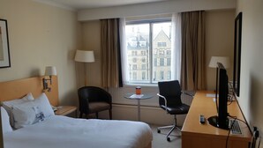 Hotellihuone, jossa on sänky, työpöytä tietokoneella, tuoli, pieni pöytä ja ikkuna, jossa on verhot.