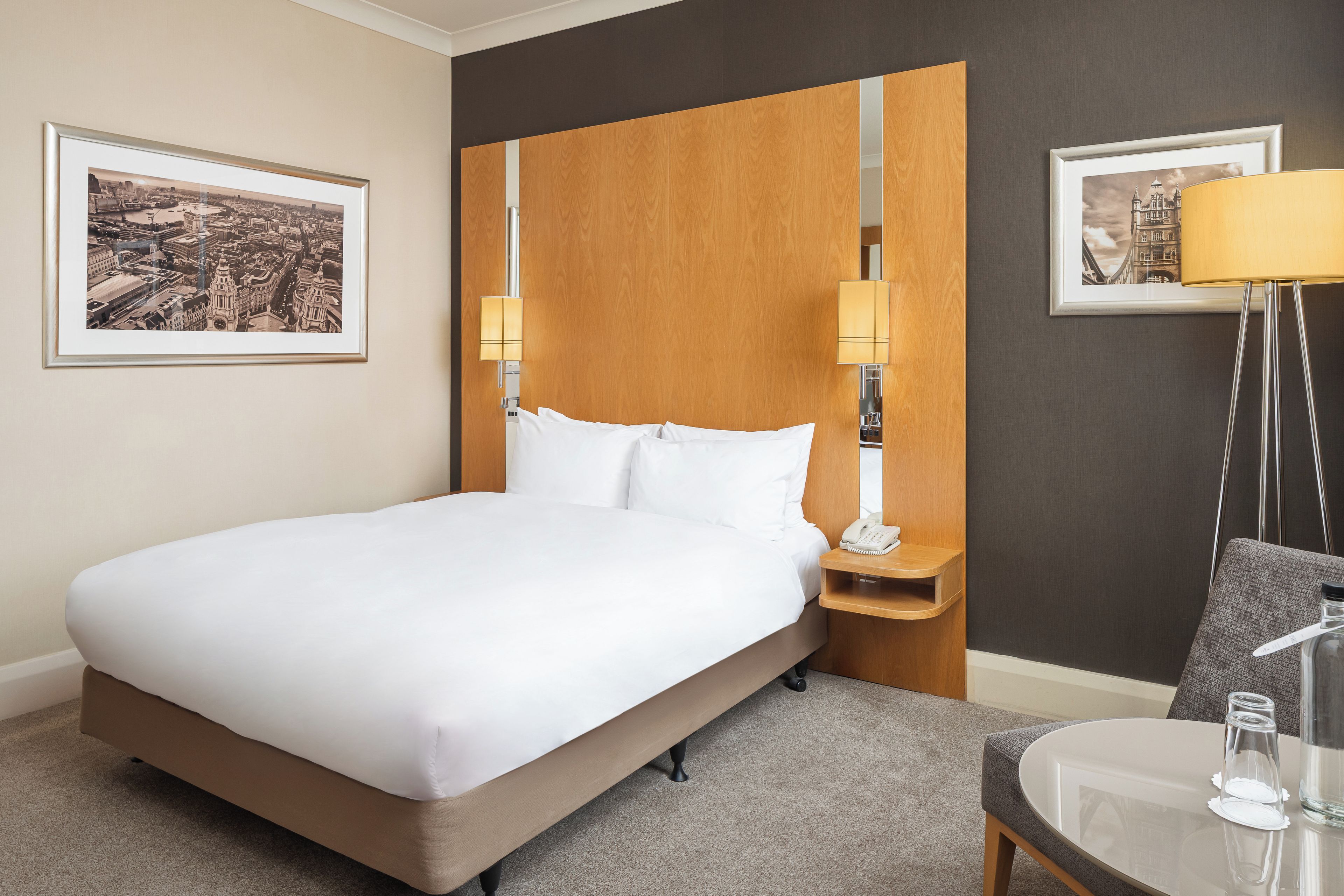 Habitación de hotel con una cama grande, una mesita de noche con lámpara, una silla y dos cuadros en la pared.