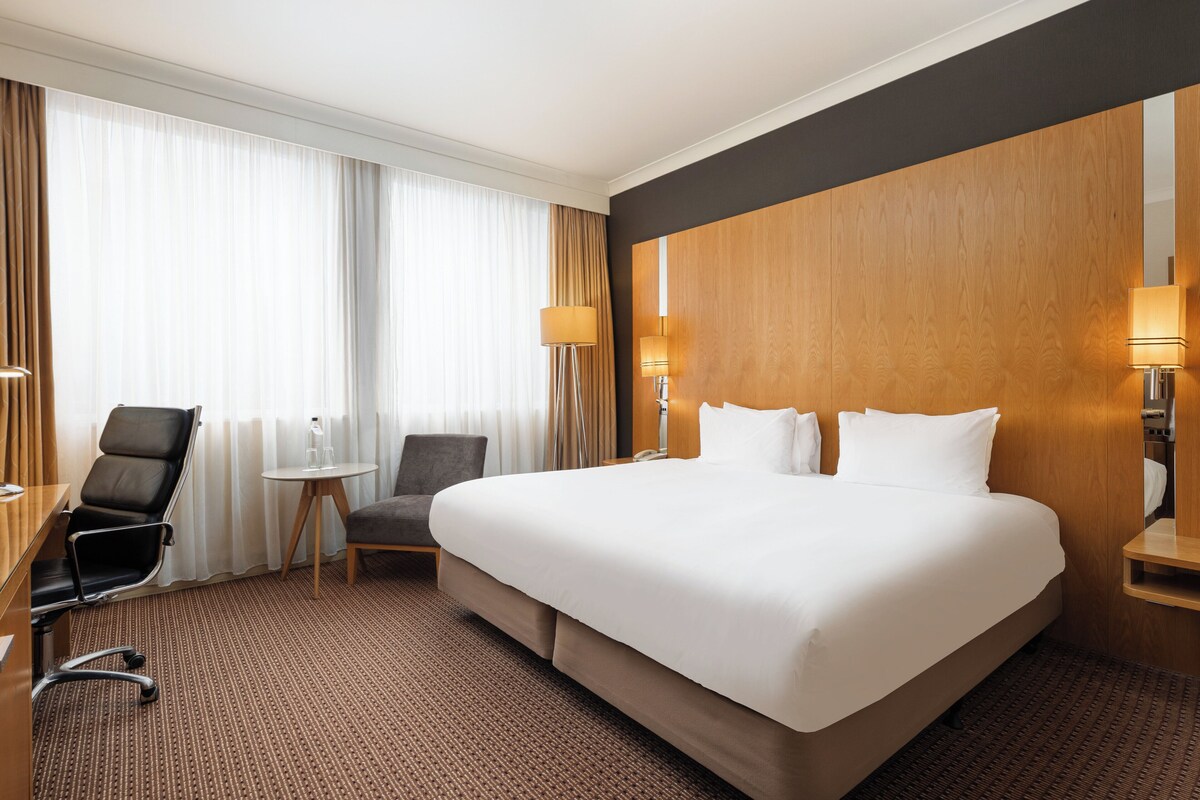 A hotel room with a large bed, a desk with a chair, a small table, and a lamp.
