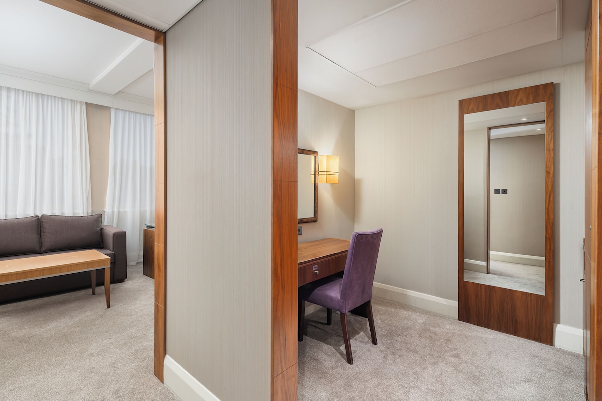 Ein Hotelzimmer mit einem Holztisch, einem violetten Sessel, einem Spiegel, einer Couch und einem großen Fenster mit Vorhängen.