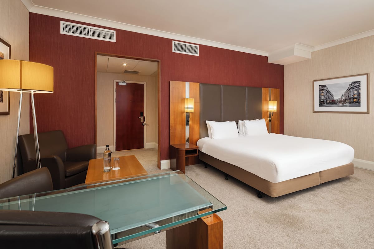 A hotel room with a large bed, a glass-top table, a desk with a chair, a lamp, and a framed picture on the wall.