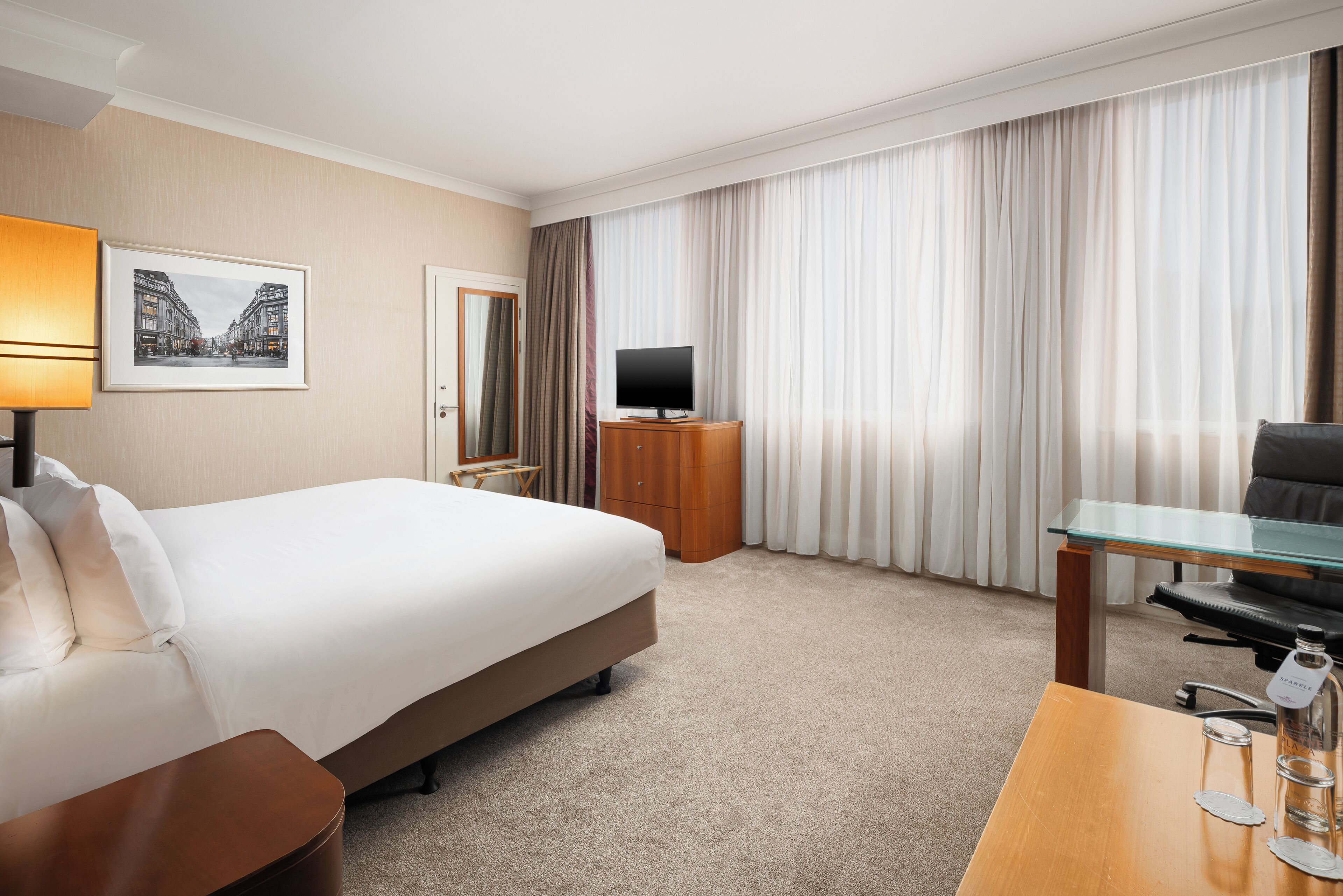 A hotel room with a large bed, a desk with a chair, a television, a mirror, and a window with curtains.