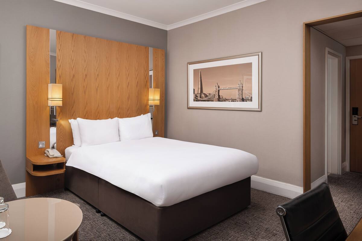 A hotel room with a large bed, wooden headboard, two bedside lamps, a framed picture on the wall, and a small table with a chair.