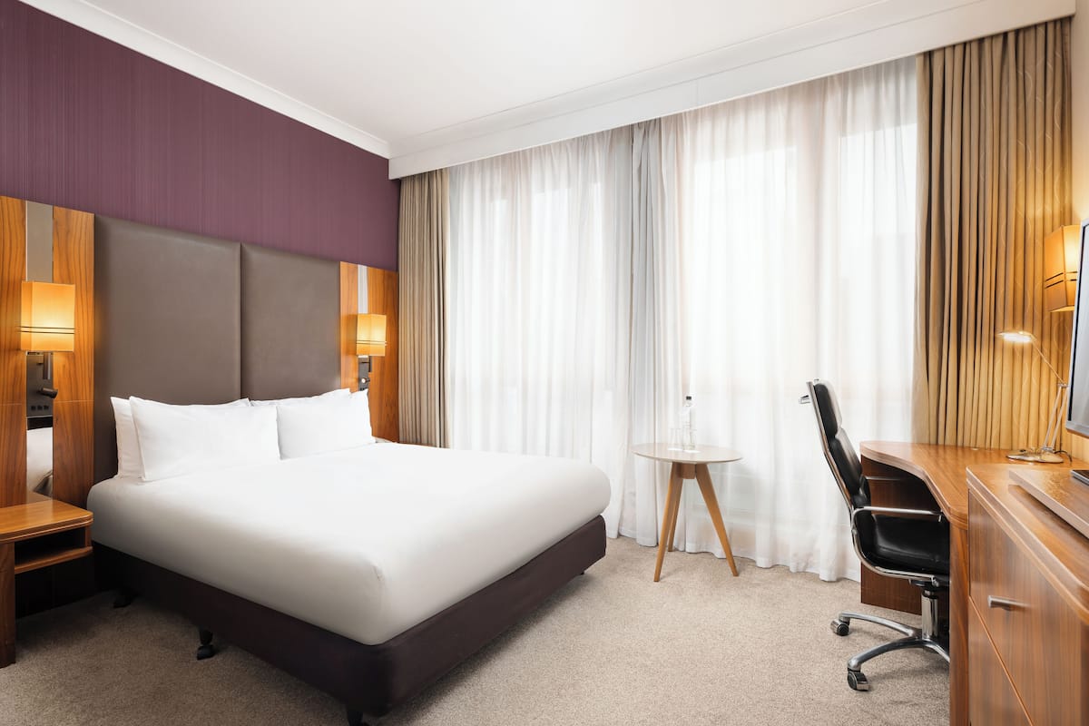 A hotel room with a large bed, a desk, a chair, a small table, and a television.