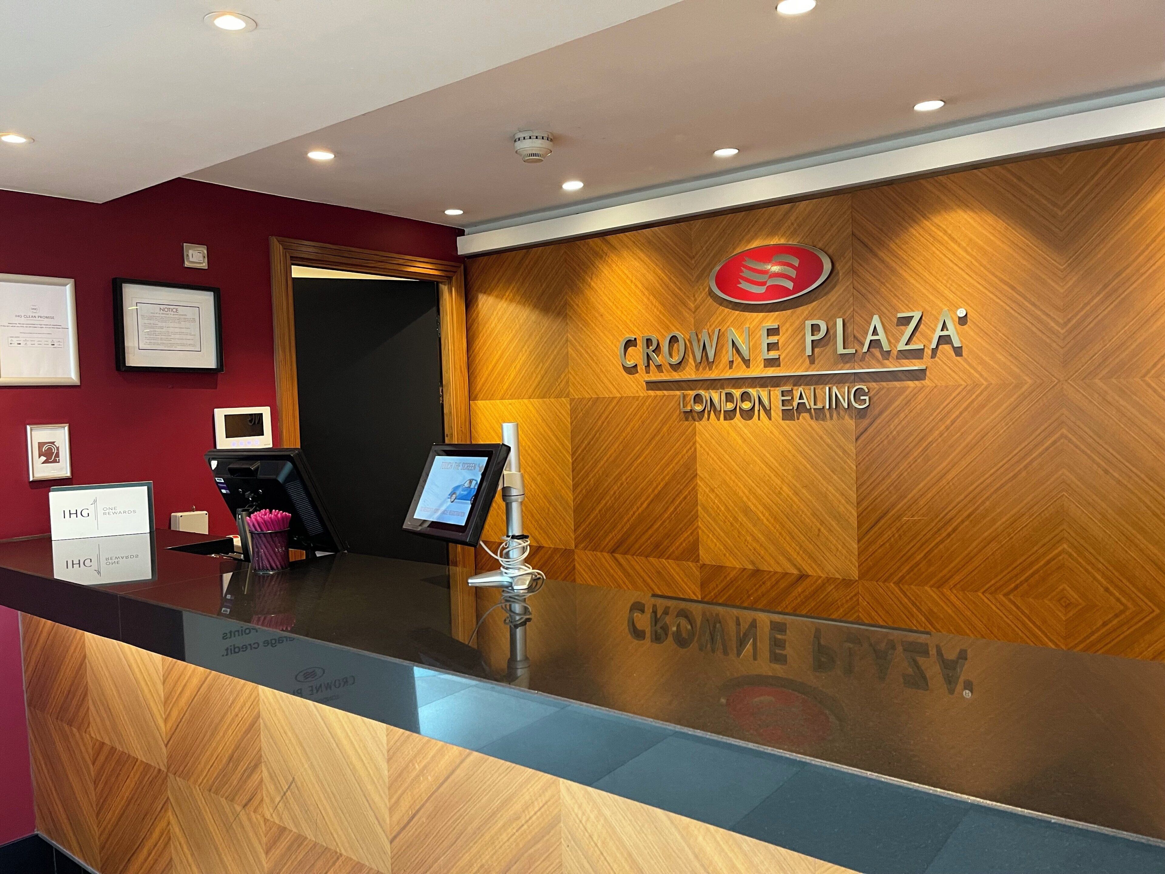 A reception desk with a digital screen, framed certificates, and a wooden wall with the Crowne Plaza London Ealing logo.