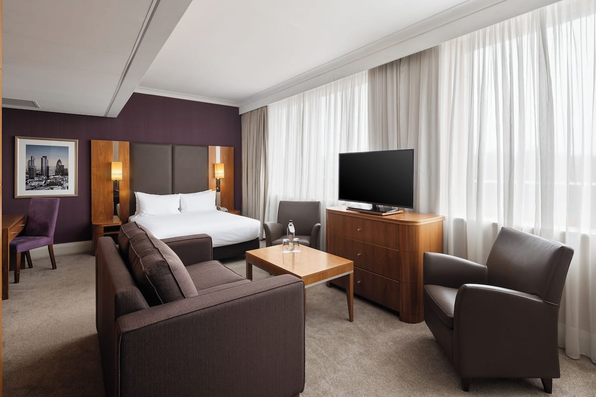 A modern hotel room with a large bed, a sofa, a desk, a TV, and a view of the city through sheer curtains.