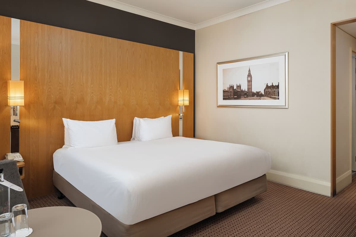 A hotel room with a large bed, two bedside lamps, a round table with a glass, and a framed picture of a cityscape on the wall.