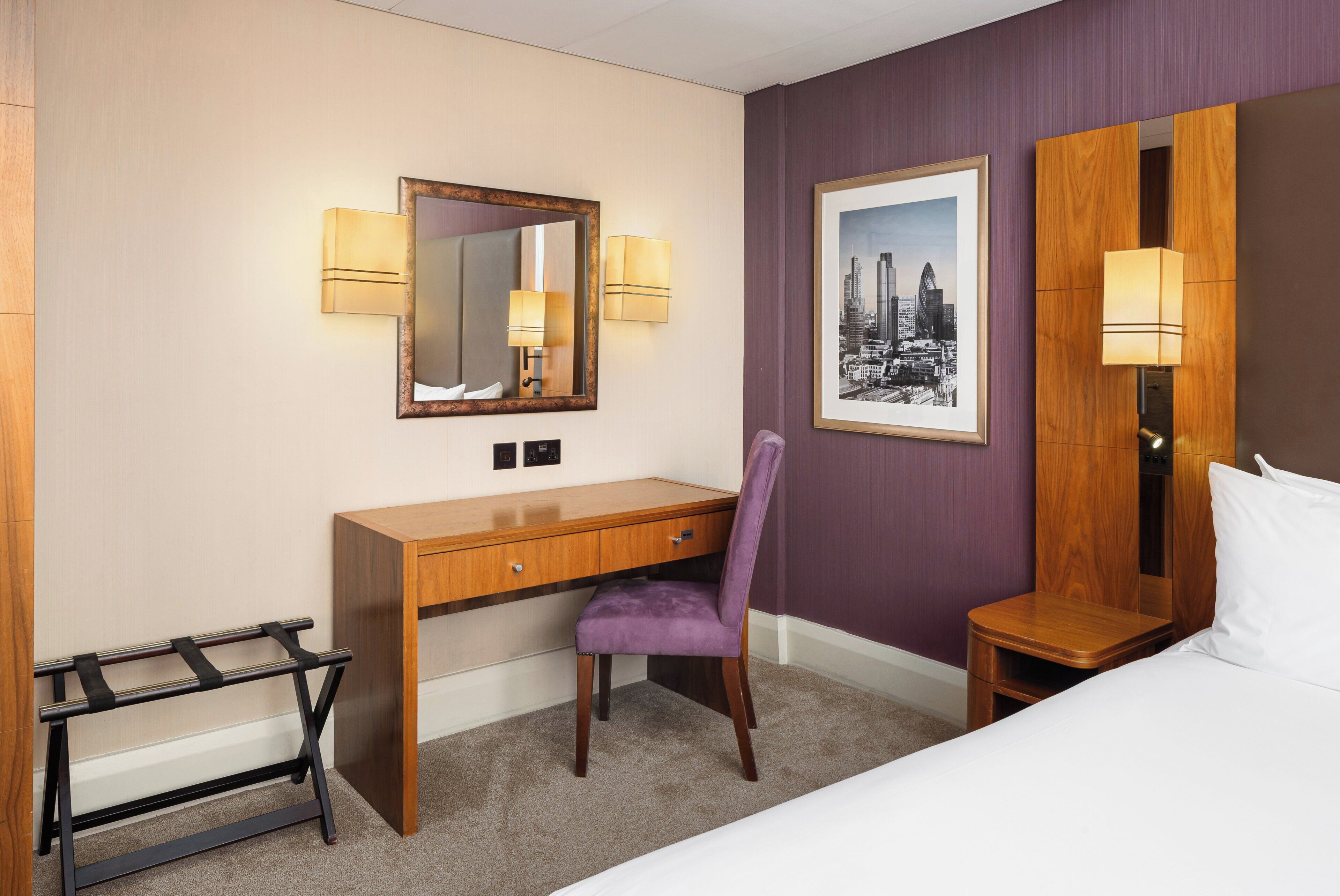 Una camera d'albergo con una scrivania in legno, una sedia viola e un letto.