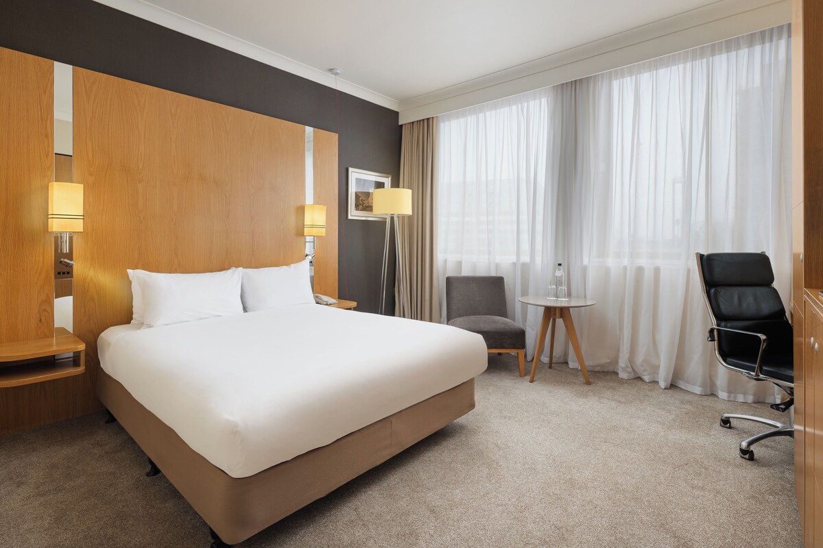 A hotel room with a large bed, a chair, a small table, and a desk with a lamp.