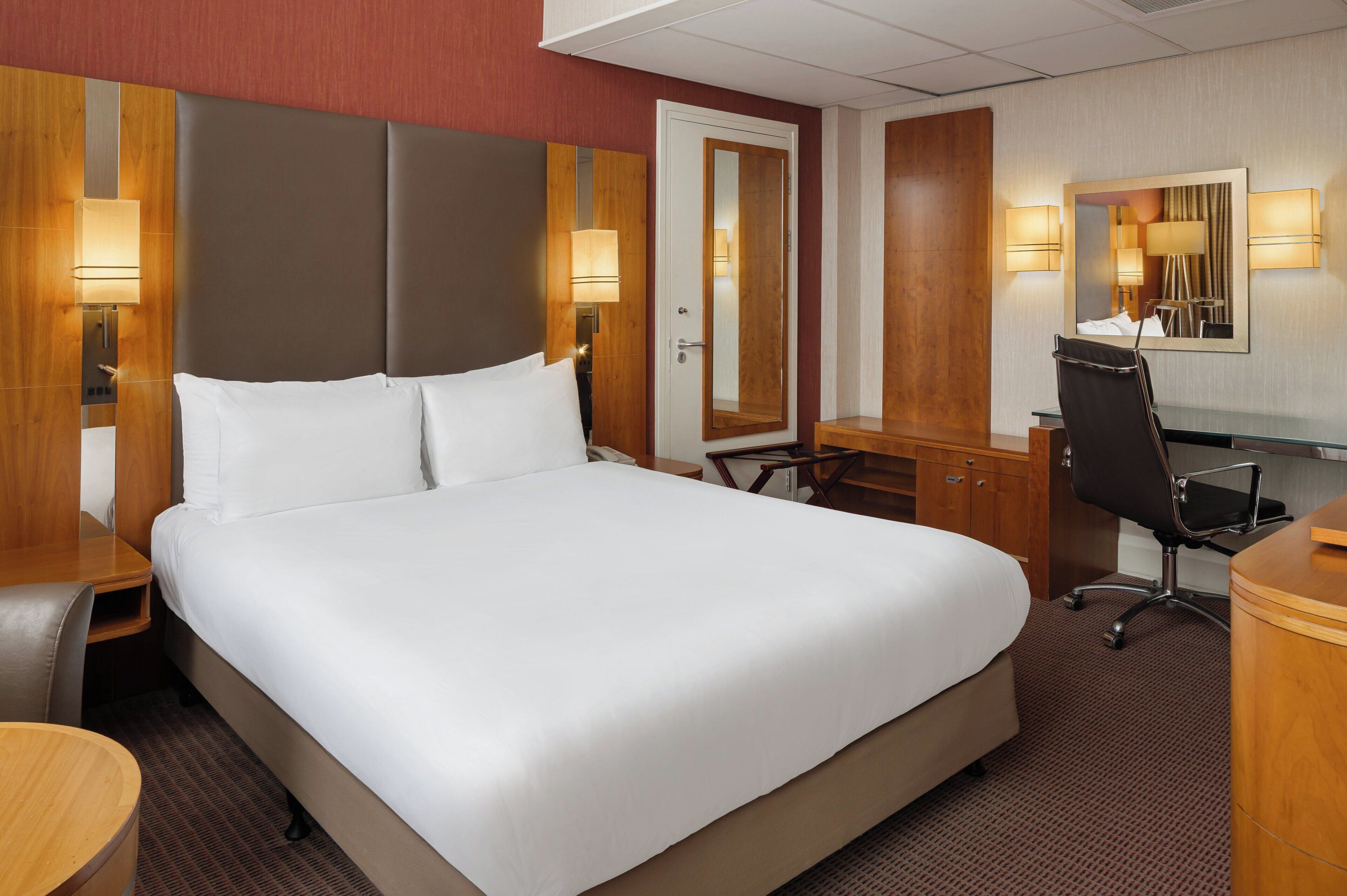 Une chambre d’hôtel avec un grand lit, un bureau, une chaise, un miroir et deux lampes fixées au mur.