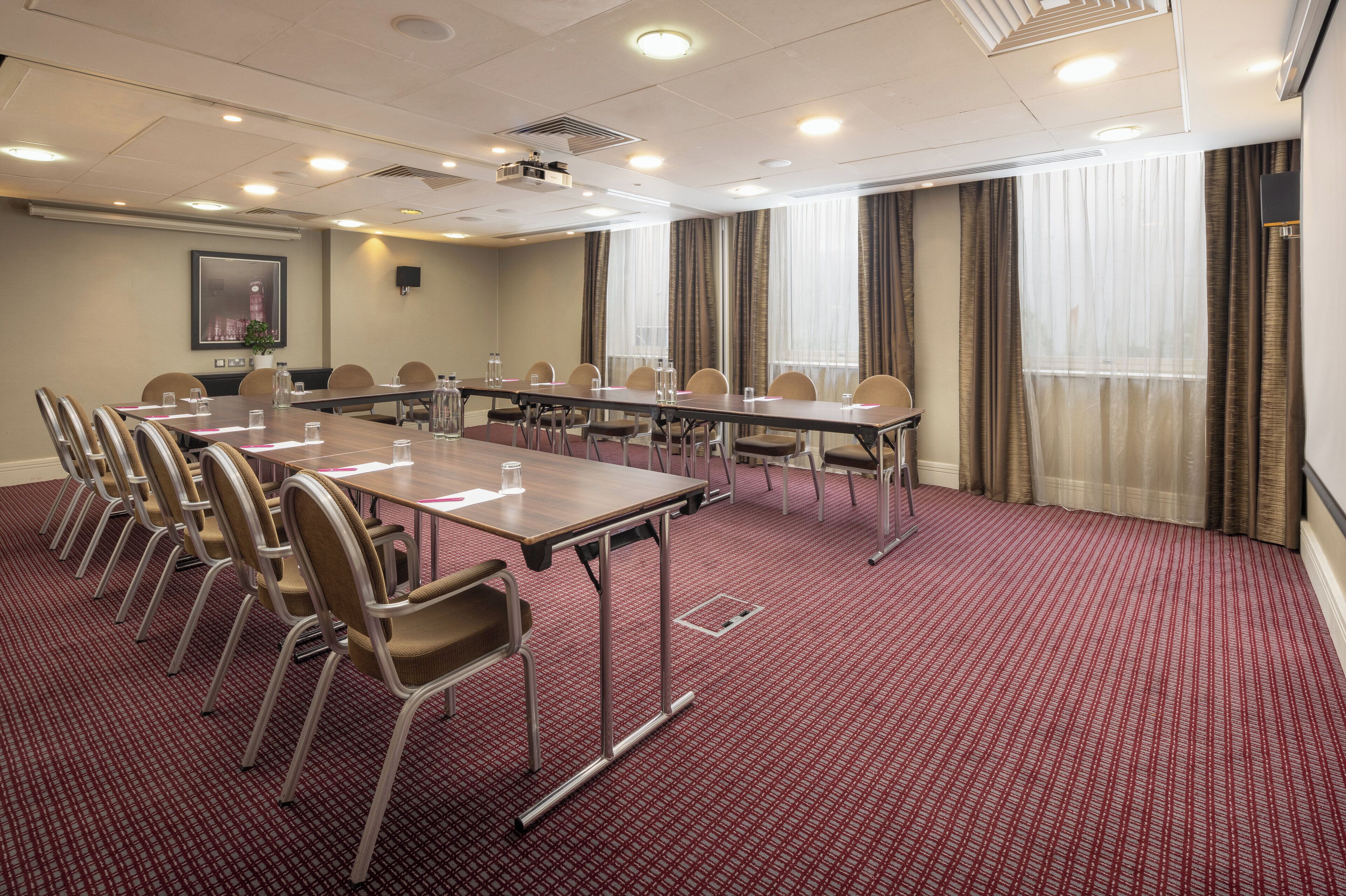 Ein Konferenzraum mit langem Tisch, Stühlen, einem Projektor und einem großen Fenster mit Vorhängen.