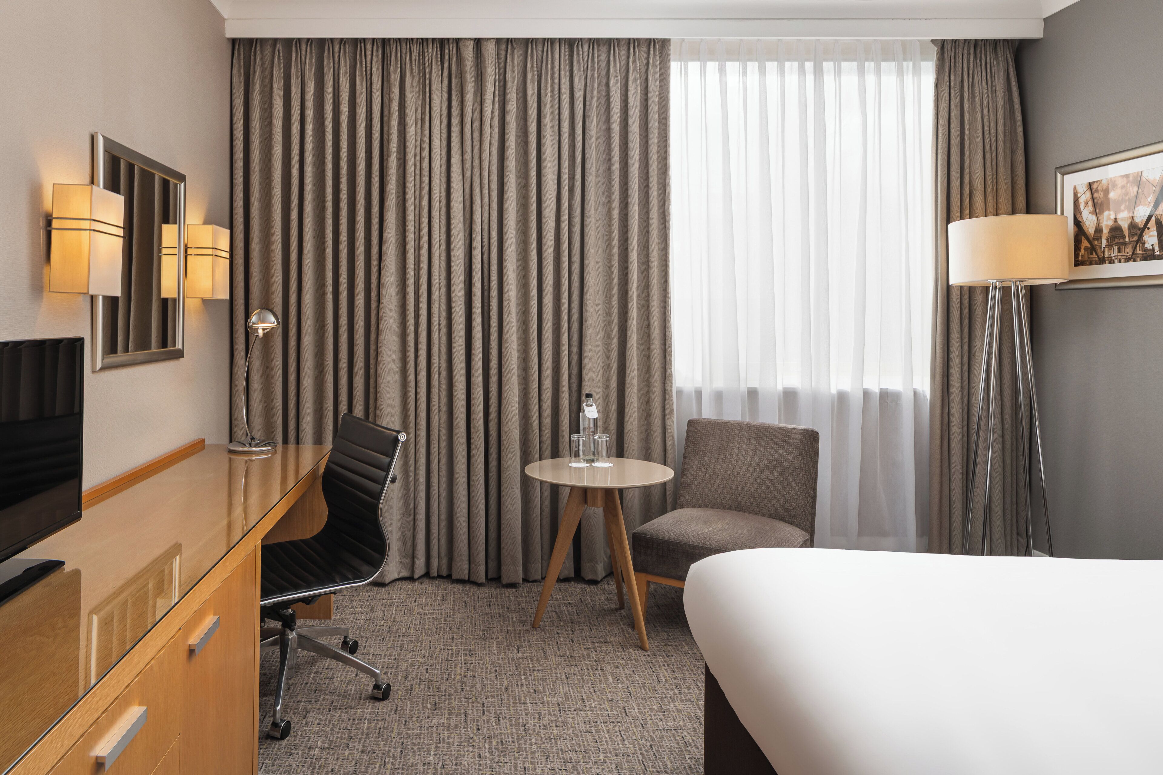 A hotel room with a bed, a desk with a chair, a round table with a chair, a TV, and a lamp.